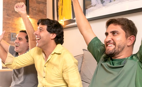 El editor especial Enzo y sus dos amigos hombres sentados en el sofá de la sala de estar, animándose con los brazos en alto y puños cerrados.