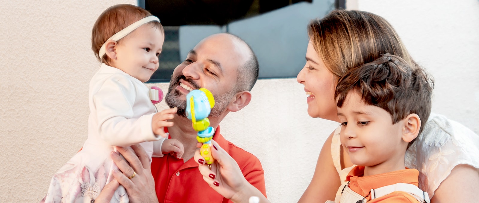 Un corte familiar, la madre le da a su hija un juguete, el padre sostiene a su hija sonriendo. Hijo mirando hacia abajo con placer en su cara.