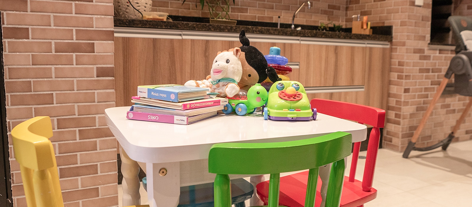 Una imagen de mesa blanca y sillas coloridas con amarillo, verde, rojo. En la mesa hay muñecas, libros y algunos juguetes para los niños.