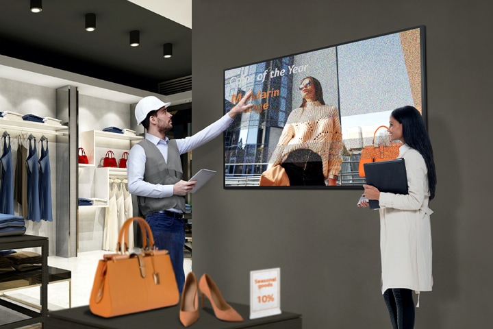 A retail store employee in a vest and hard hat gestures toward a digital display showing a woman with "Employee of the Year" text. Another person, holding a folder, observes the screen in a modern retail space with shelves of clothing and bags.