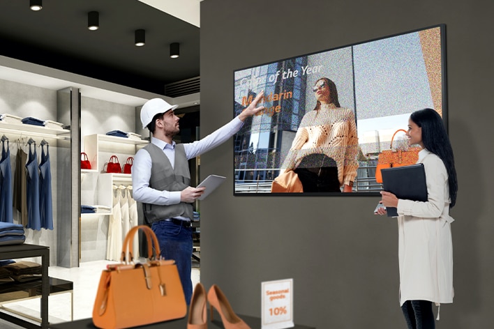 A retail store employee in a vest and hard hat gestures toward a digital display showing a woman with "Employee of the Year" text. Another person, holding a folder, observes the screen in a modern retail space with shelves of clothing and bags.