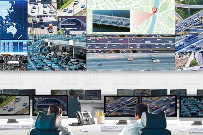 A traffic control room with multiple monitors displaying real-time footage of highways, traffic maps, and analytics. Two operators monitor the screens, showcasing advanced traffic management and surveillance systems.