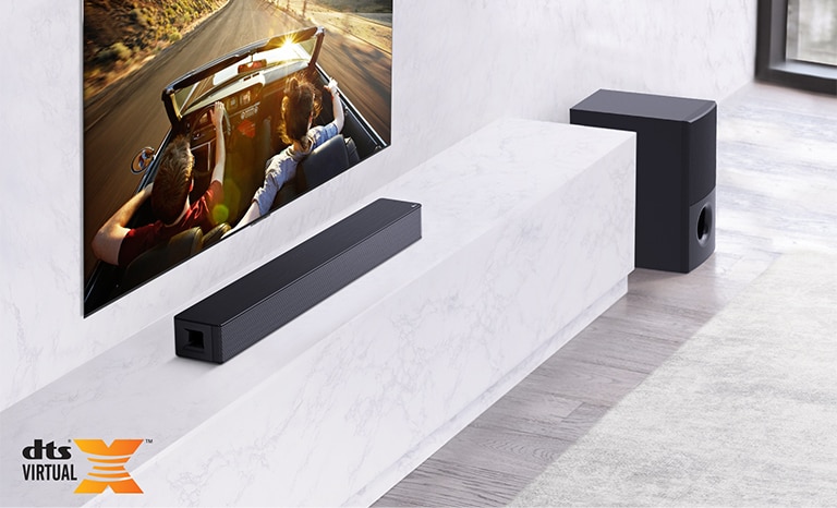 TV is on the wall, LG Soundbar is below on a white marble shelf with a sub-woofer to the right. TV shows a couple in a car.