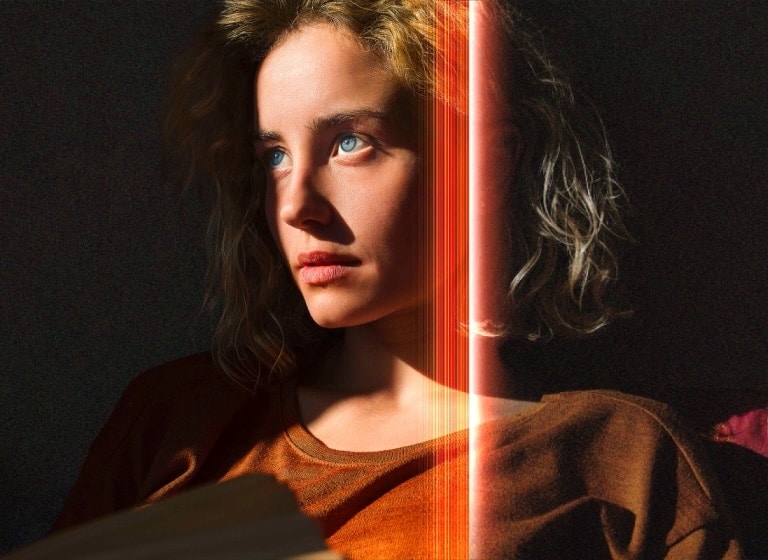 An video of a woman with piercing blue eyes and a burnt orange top in a dark space. Red lines depicting AI refinements cover part of her face, which is bright and detailed, while the rest of the image looks dull.