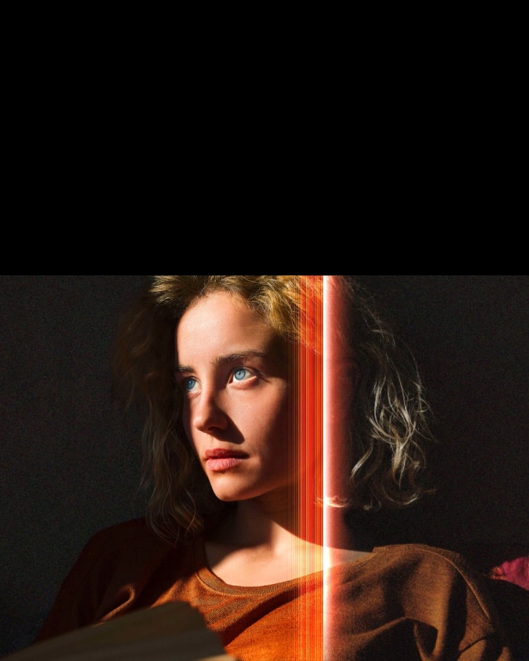 An video of a woman with piercing blue eyes and a burnt orange top in a dark space. Red lines depicting AI refinements cover part of her face, which is bright and detailed, while the rest of the image looks dull.	