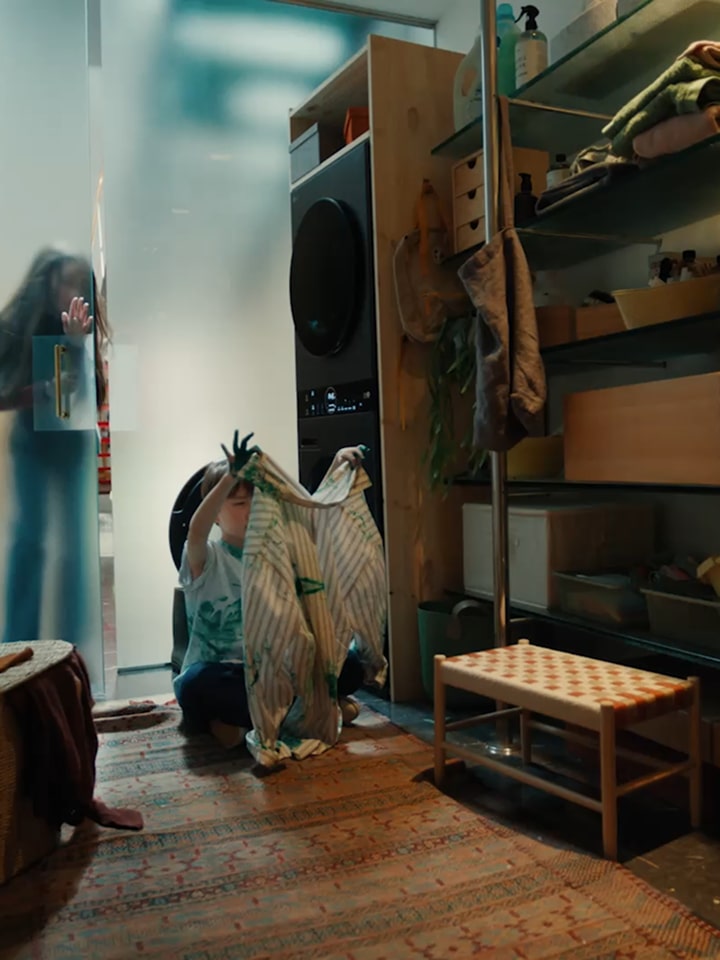 A woman slides open a frosted glass door and enters. Inside the laundry room, a boy is placing laundry into the LG washing machine.The washing machine screen displays the text &quot;AI Wash.