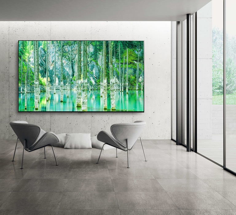 Beeld van een grootbeeld LG QNED MiniLED TV gemonteerd tegen een grijze muur. Twee stoelen staan voor de tv en het scherm toont een groen natuurtafereel.