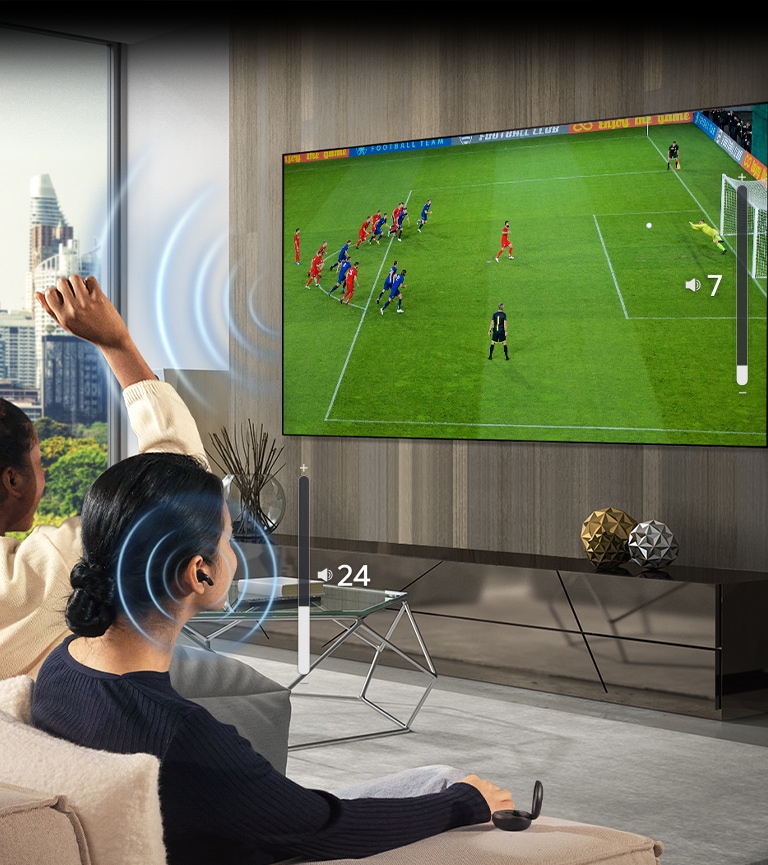 Een groep mensen zit op een bank naar een voetbalwedstrijd op tv te kijken. De vrouw uiterst rechts draagt earbuds en gebruikt die met een ander volume dan de tv, wat erop wijst dat zij beide tegelijk gebruikt.