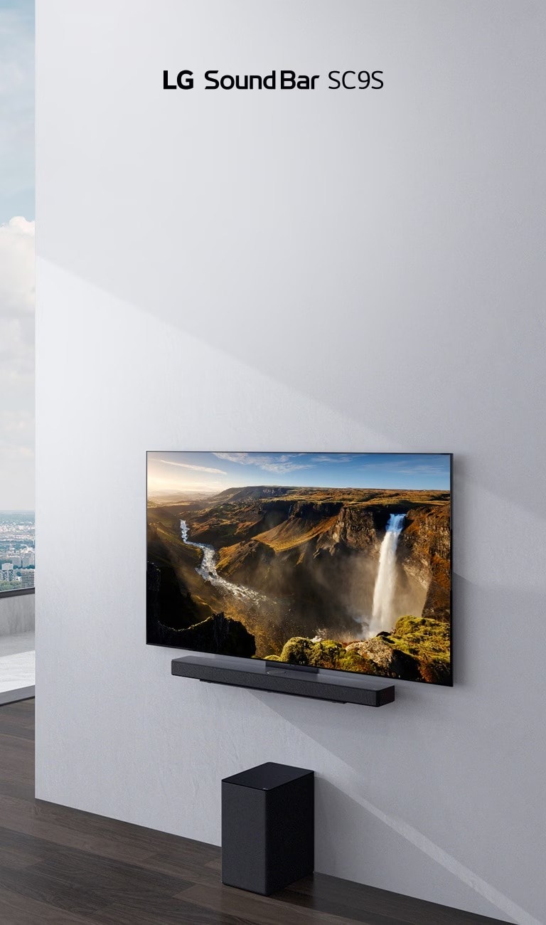 TV and LG Sound Bar SC9S are hung on a white wall. Below a black wireless subwoofer is placed on the floor. Beyond the window on the left a city view with the blue sky can be found.