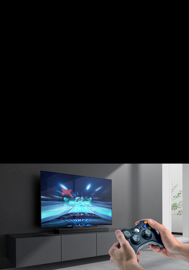 The sound bar is placed on the cabinet and racing game scene is shown on the TV coneected to the sound bar. A game console is on the bottom right side of the picture hold by two hands.