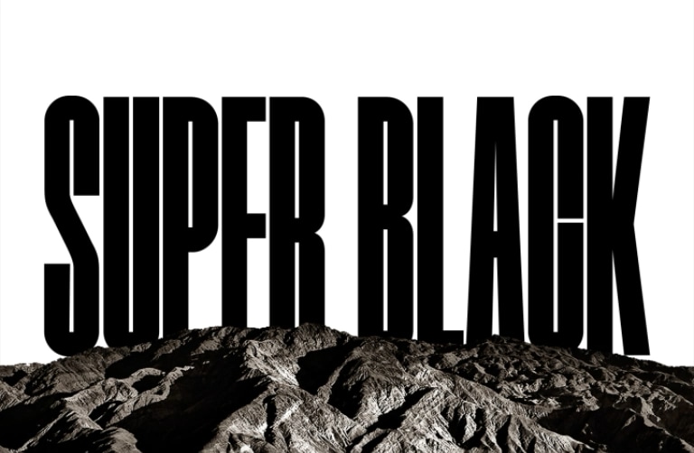 A video opens with the words "SUPER BLACK" in bold black capitals. A black mountainous scene with crisp definition then rises to cover the letters, also revealing a village and sand dunes. The black copy disappears behind a black sky.