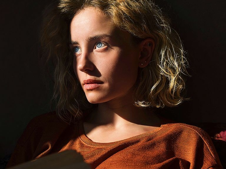 An video of a woman with piercing blue eyes and a burnt orange top in a dark space. Red lines depicting AI refinements cover part of her face, which is bright and detailed, while the rest of the image looks dull.