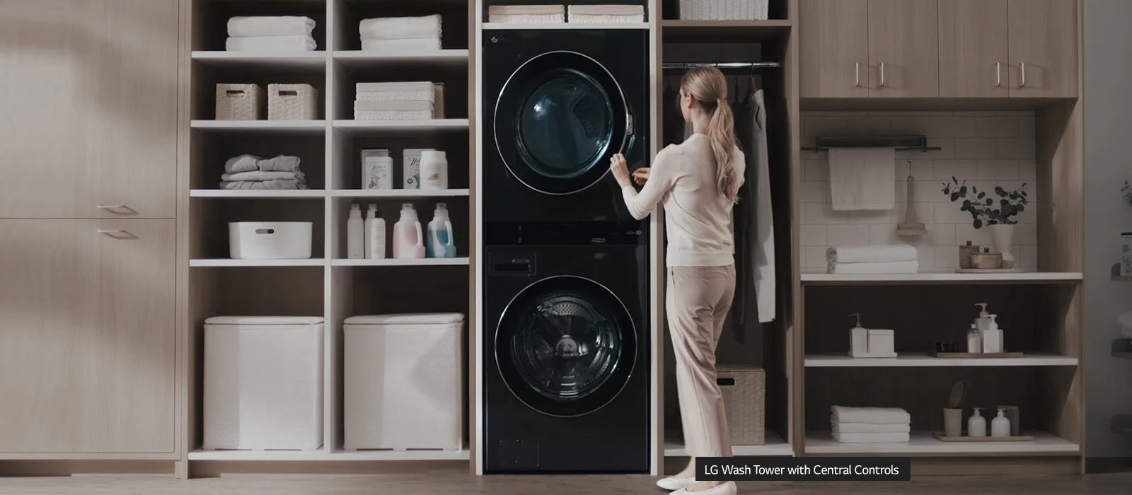 A white washer and dryer are stacked on top of each other and installed in a wall unit with shelves and a closet and a woman has her back facing the front as she reaches up to press a button on the center top control panel. She stands uncomfortably.