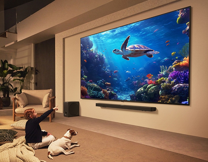 Una enorme LG QNED TV y LG Soundbar montadas en la pared. Una niña y su perrito están mirando una escena bajo el agua en la TV. El gran tamaño de la TV hace que el contenido se sienta aún más inmersivo.