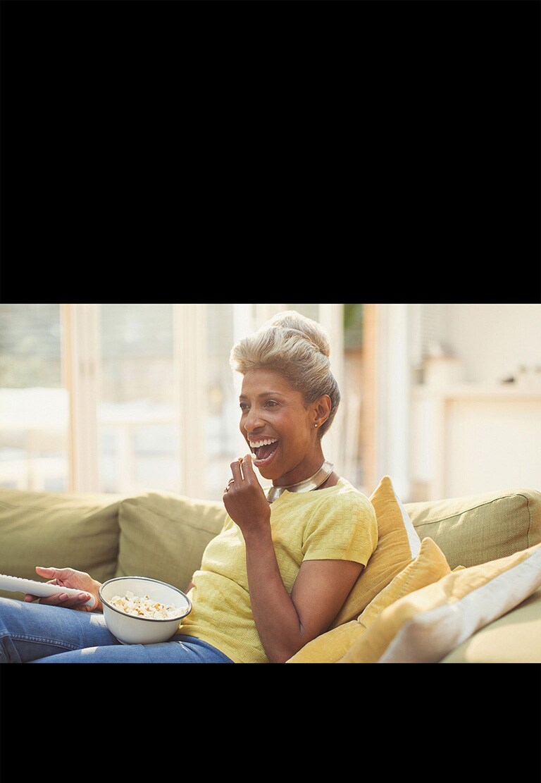 Una mujer está viendo la televisión, y sostiene un control remoto. También está comiendo palomitas de maíz.
