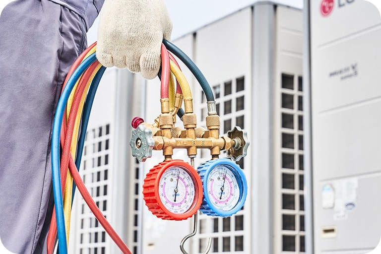 In the background, there are large LG HVAC units, and in front of them, a technician's gloved hand holds a set of HVAC manifold gauges with colorful hoses used for measuring refrigerant pressure.