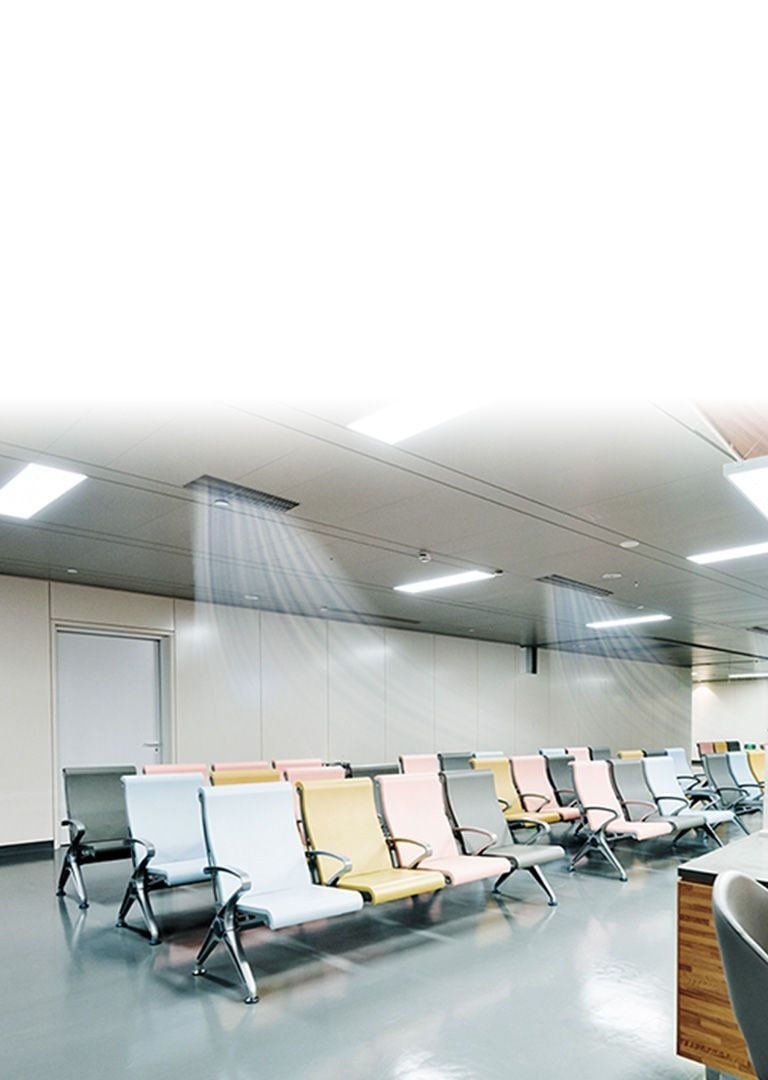 Three ceiling mounted air conditions create blue-colored air stream in a waiting room in hospital with a series of chairs and a reception desk to the right.