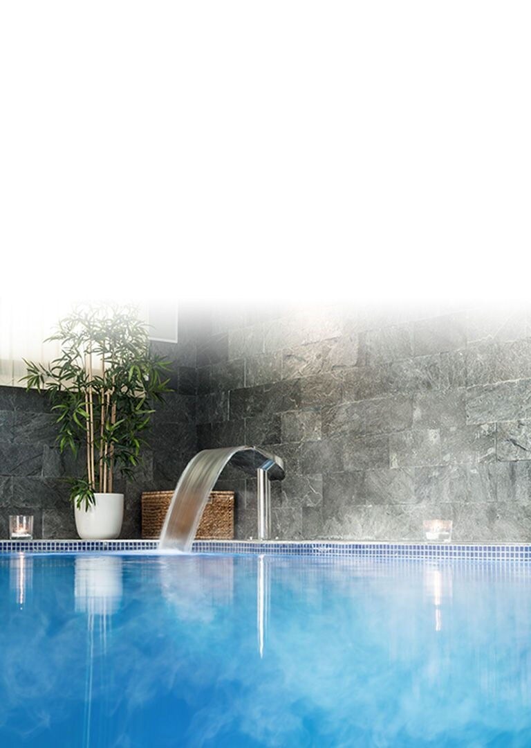 An indoor swimming pool with walls coverd with grey marble square tiles and an artificial waterfall to the left.