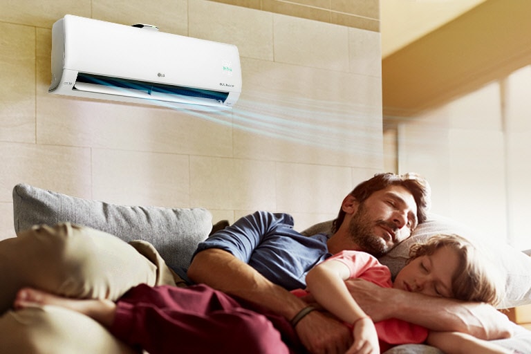 A father and daughter sleep on a couch beneath an air conditioner that is blowing air out over them.