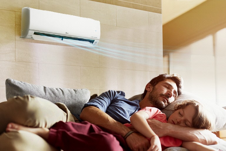 A father and daughter sleep on a couch beneath an air conditioner that is blowing air out over them.