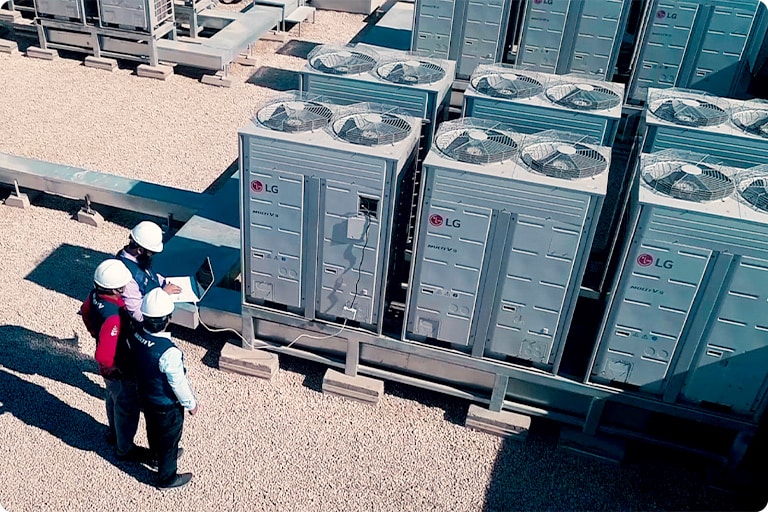 Three helmeted people peer at a laptop screen amidst an area with a group of LG outdoor units neatly installed.