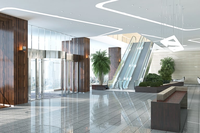 Building lobby with revolving door at entrance. Reception desk straight ahead. Escalator on right surrounded by plants.