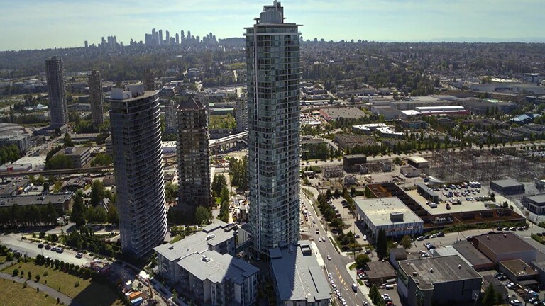 In daylight, a mega city view reveals a tall, round glass building at the center, flanked by shorter buildings to the left, under a cloudy blue sky.