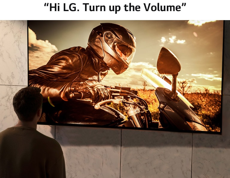 The back of a man watching TV on the wall in the living room. There is a motorbike movie scene on the screen.