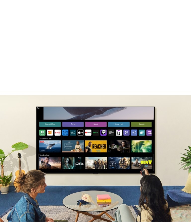 A whale floating above the ocean with a woman in the ocean are shown. A home screen appears from below. As the scene transitions, it shows the image in a large LG TV on the wall. Two women sit in a cozy and neutral living room filled with plants, and a guitar. One woman points the remote at the TV, which shows a range of apps and recommended TV shows on the home screen.