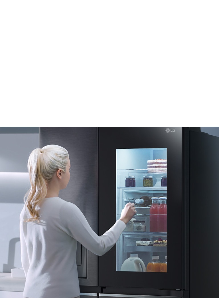 A video shows a woman approach her InstaView refrigerator and knock twice. The interior lights up and she can see the contents of her fridge without opening the door. The view zooms in to focus on the drinks in the door and then zooms out to see the woman from behind as she opens the door and grabs a drink.