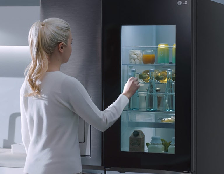 A video shows a woman approach her InstaView refrigerator and knock twice. The interior lights up and she can see the contents of her fridge without opening the door. The view zooms in to focus on the drinks in the door and then zooms out to see the woman from behind as she opens the door and grabs a drink.