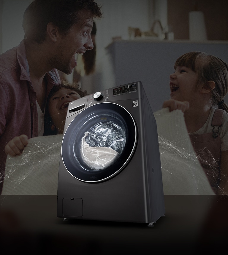 Father and daughters laugh in the background as they hold a clean blanket. A White washing machine front load washer in the foreground.
