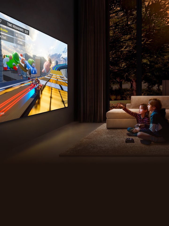 A side-angle view of an ultra-large LG TV mounted on a wall in a cozy living room displaying a colorful game, and two children sitting on the floor in front of the TV holding game controllers.