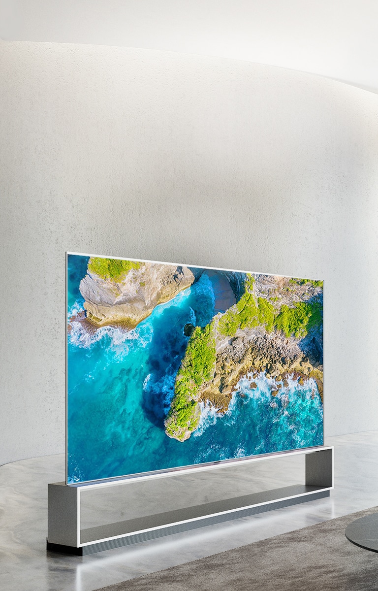 Telewizor LG SIGNATURE OLED W9 jest zawieszony na ścianie, a przed telewizorem leży kranik z niebieskim niebem nad oknem.
