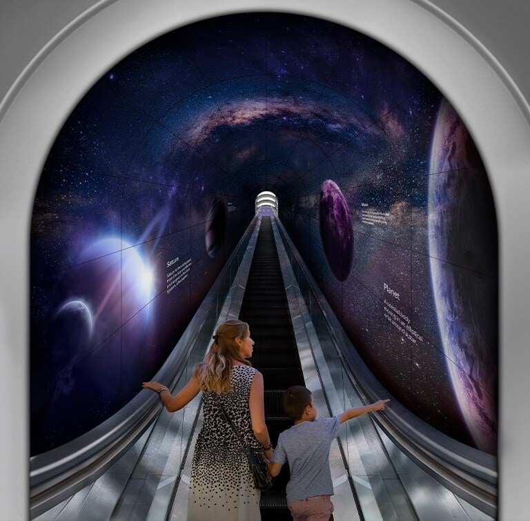 Kobieta z dzieckiem idą tunelem wykonanym z elastycznej zakrzywionej ramy ekranu OLED Signage po schodach ruchomych.