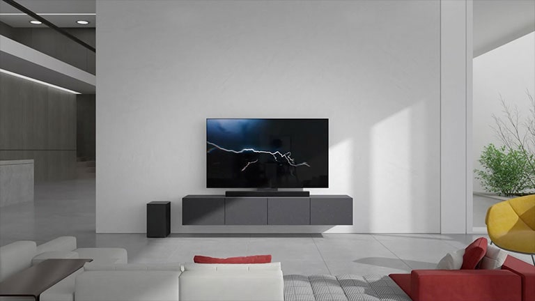 Soundbar stoi na szarej szafce z telewizorem w pokoju dziennym. Czarny bezprzewodowy subwoofer stoi na podłodze po lewej stronie, a z prawej strony obrazu dochodzi światło słoneczne. Biało-czerwona długa kanapa stoi naprzeciwko telewizora i soundbaru.