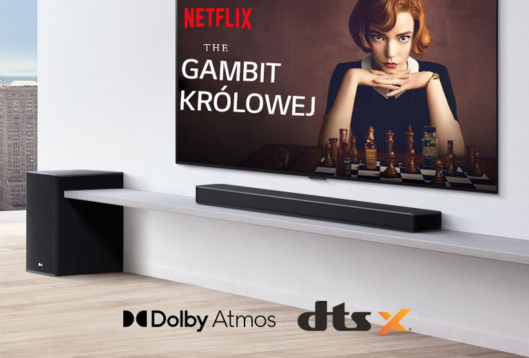 Telewizor zawieszony na ścianie.  Plakat programu telewizyjnego na ekranie. Soundbar LG znajduje się bezpośrednio pod telewizorem, na białej półce, tuż obok stoi subwoofer. Logo Dolby Atmos i DTS:X są wyśrodkowane u dołu ekranu.