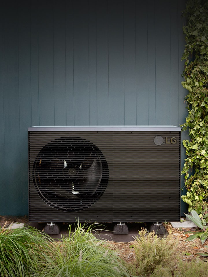 LG Air to Water Heat Pump THERMA V R290 Monobloc, black-colored outdoor unit is placed on the exterior green wall of the house.