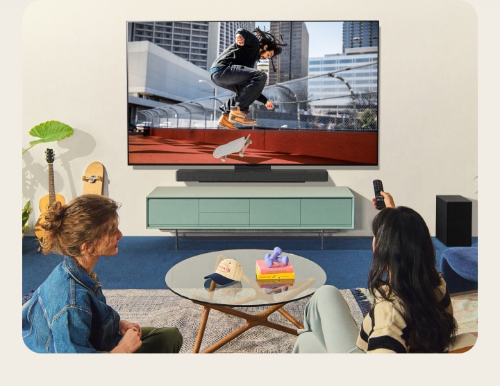 Duas mulheres sentadas num sofá em frente a uma LG TV montada na parede numa sala de estar colorida, e uma mulher aponta um controlo remoto para o ecrã da TV. 