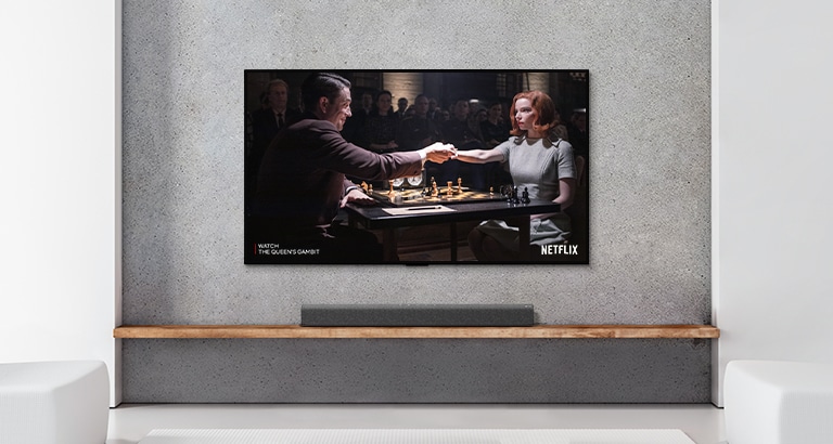 Uma barra de som e TV numa sala branca. Um casal joga xadrez no ecrã da TV.