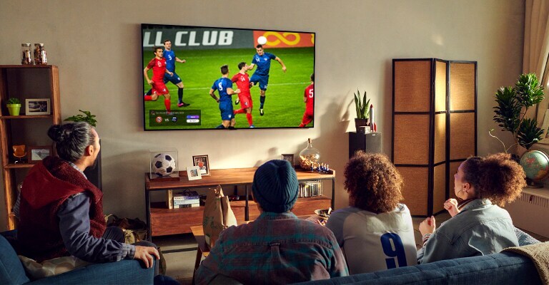 Um homem e uma mulher sentados numa mesa de café à frente de uma TV montada na parede com um jogo de futebol.