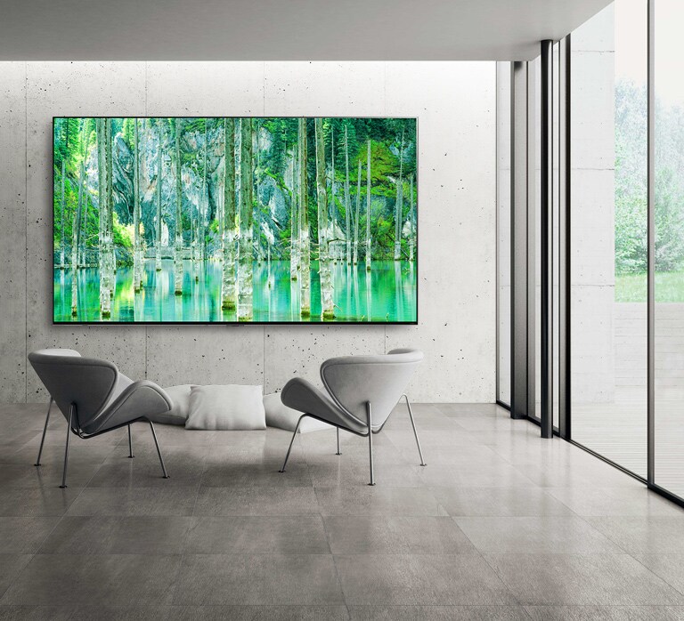 Imagem de uma TV LG QNED MiniLED do ecrã grande montado contra numa parede cinza de betão nua. Duas cadeiras estão dispostas em frente à TV e o ecrã mostra uma cena natural verde.