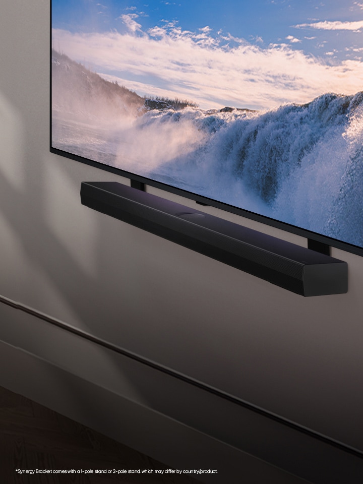 LG TV and Soundbar are placed within an angled perspective mounted on a wall. On the TV, a close up of a vast waterfall is displayed, and soft sunlight cascade over the wall, TV, and soundbar. A disclaimer reads: "Synergy Bracket comes with a 1-pole stand or 2-pole stand, which may differ by country/product."