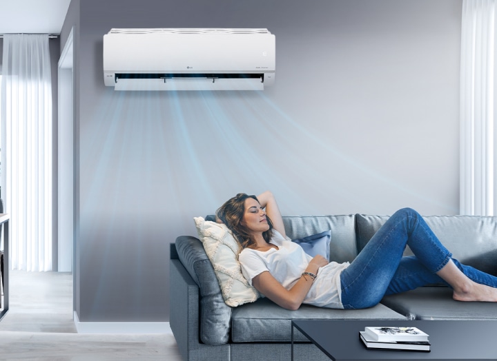 A woman lounges on a couch in a living room with the LG air conditioner installed above her on the wall. Blue streams of air are on the image to indicate it is on and cooling the room.	