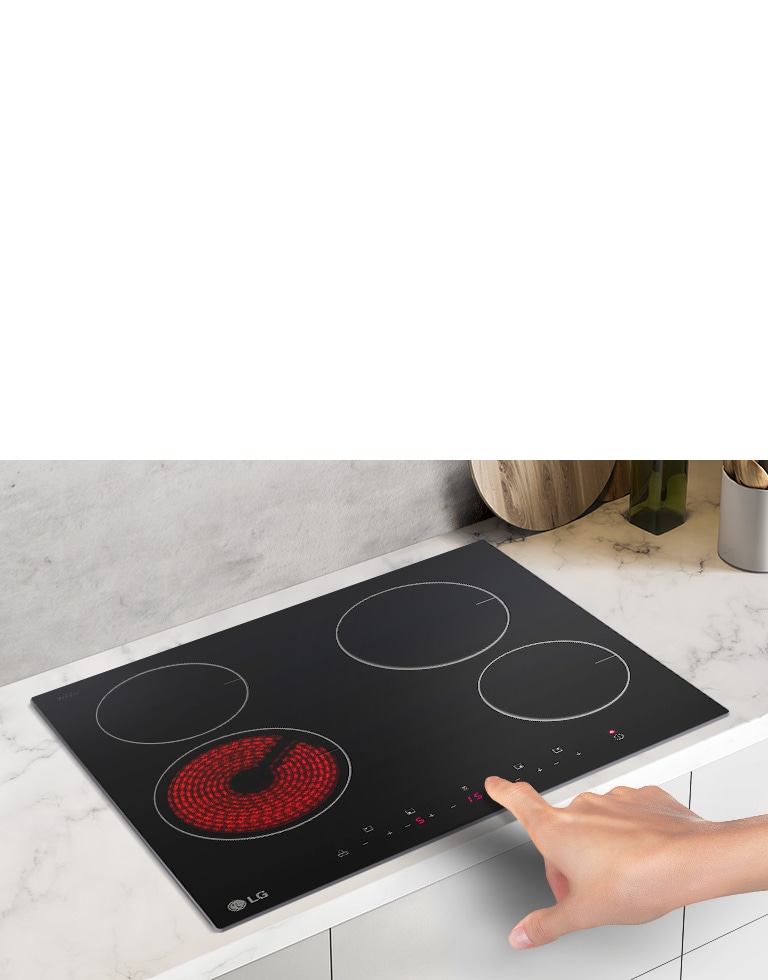 The STUDIO cooktop is seen from an angle up looking down. A hand reaches in to press a button. The left-front burner is red with heat.