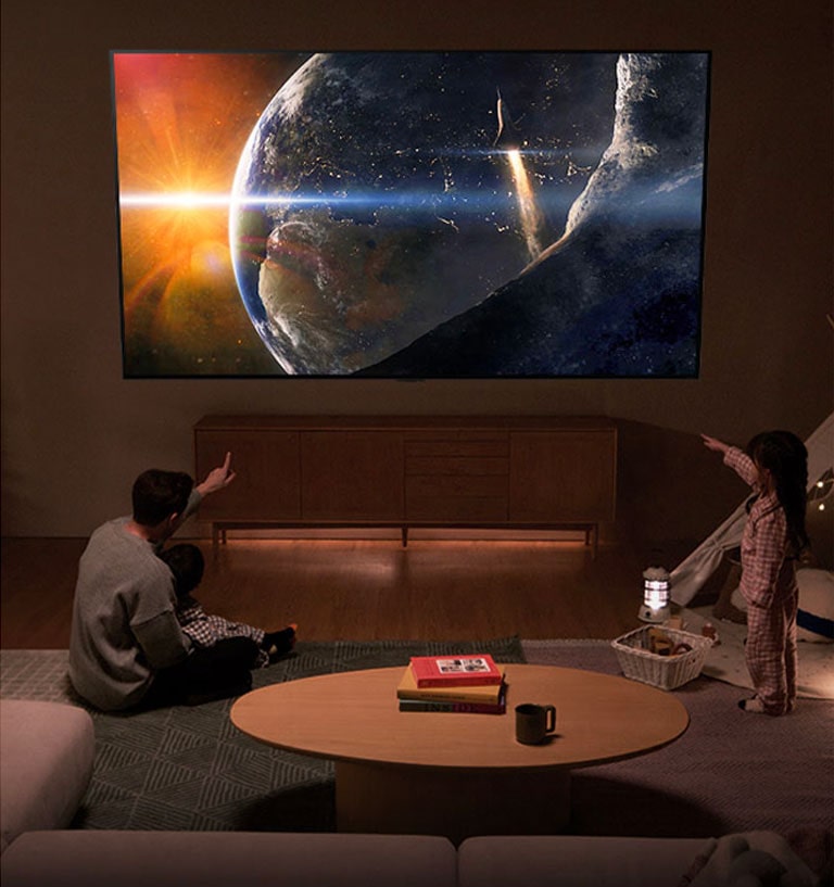 A family sat on the floor of a low-lit living room by a small table, looking up at an LG TV mounted on the wall showing the Earth from space.