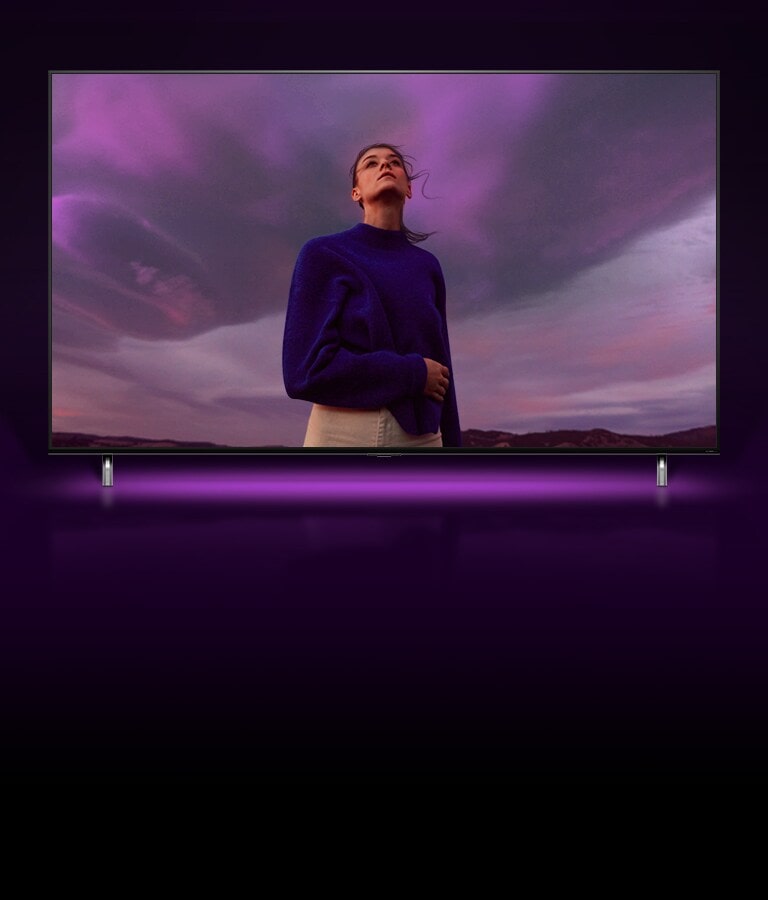 A woman is standing looking at a purple sky and the image zooms out to show the same woman in QNED TV screen.