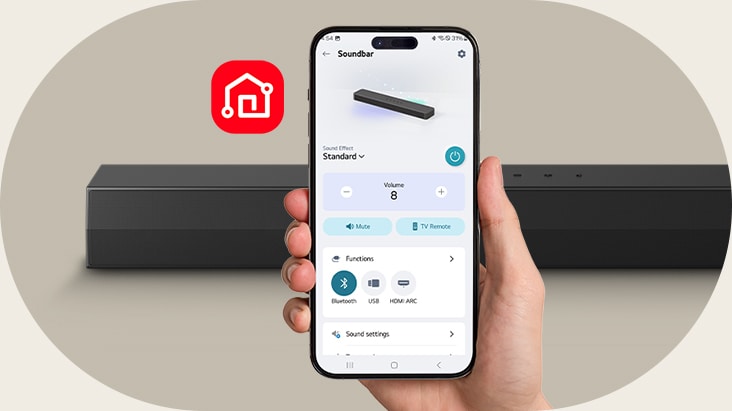 En smartphone med LG ThinQ-appen öppen hålls i en hand. Bredvid den visas LG ThinQ-appens ikon. Bakom finns en SH5A Soundbar.