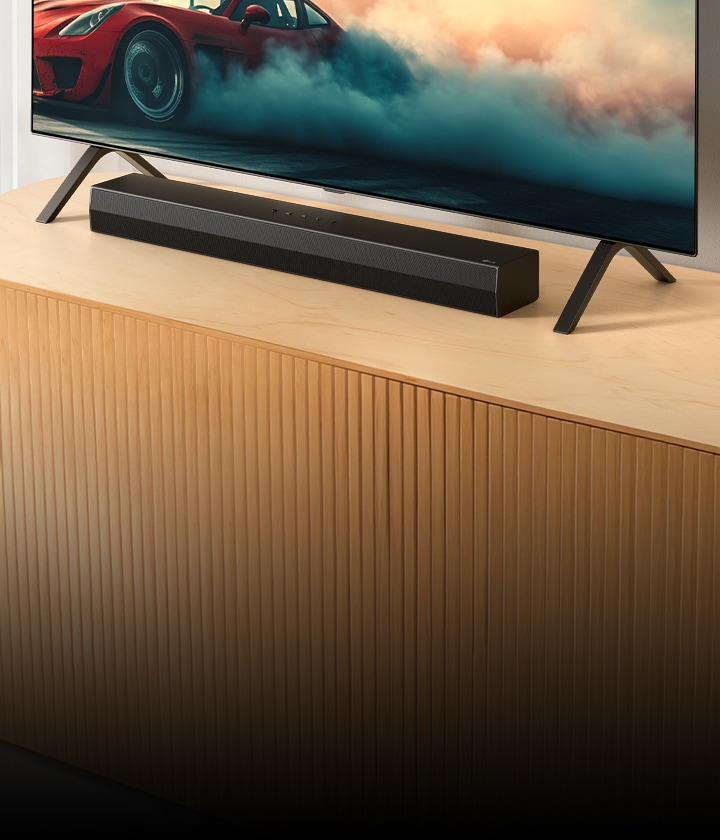 LG Soundbar S20A placeras på en TV-konsol i trä, under en TV och mellan dess TV-stativ. På TV:n visas en röd bil med rök.