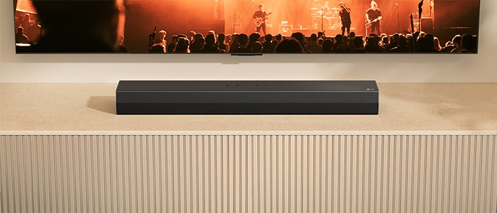 En S20A soundbar är placerad på en TV-konsol i trä. Ovanför visas en del av en TV som visar en konsertspelning med ett band.